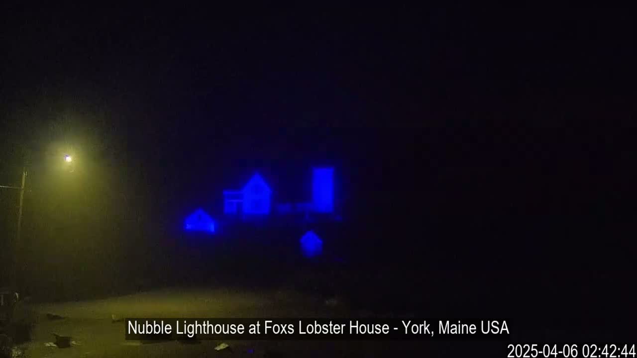A dimly lit scene in foggy conditions shows a lighthouse and outbuildings illuminated in blue.