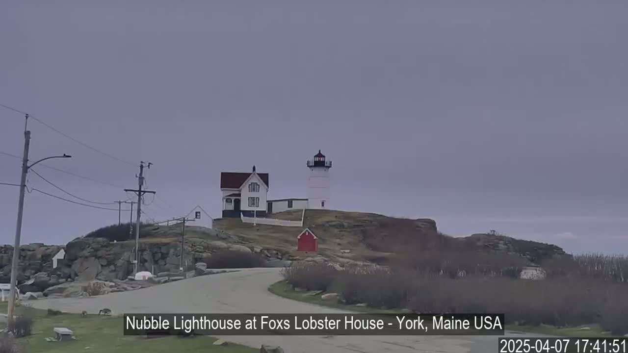 A lighthouse and accompanying buildings sit atop a rocky, sparsely vegetated hill under an overcast sky.