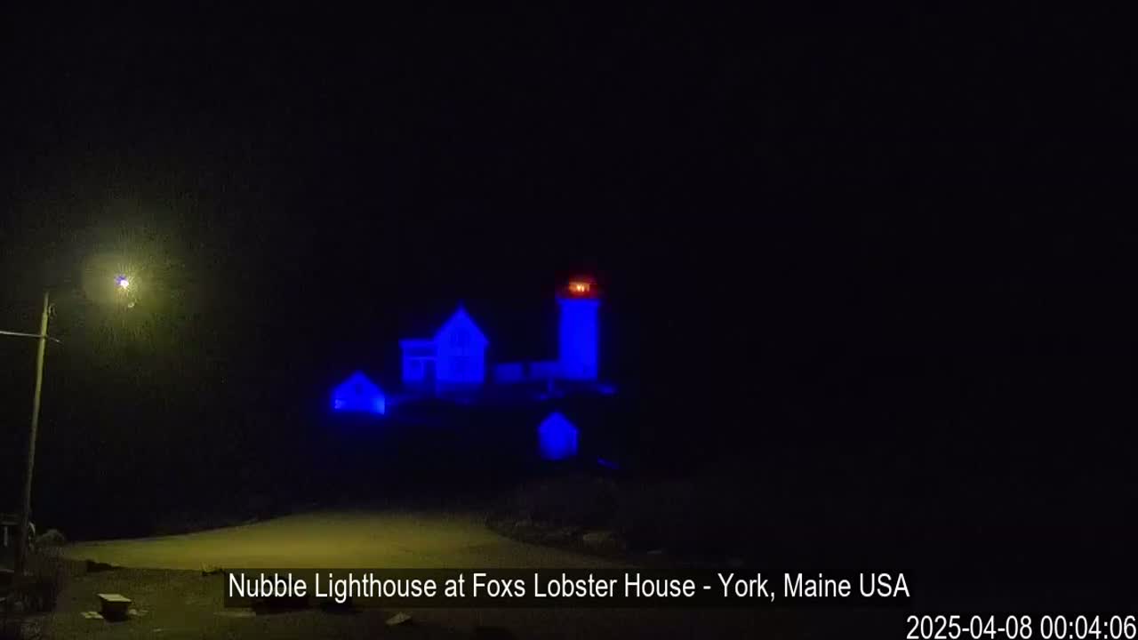 A lighthouse and nearby buildings are illuminated in blue at night under a dark sky.