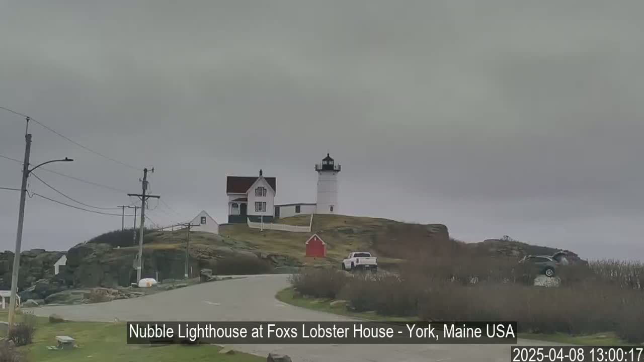 A lighthouse and a smaller house sit atop a rocky hill overlooking a road under an overcast sky.
