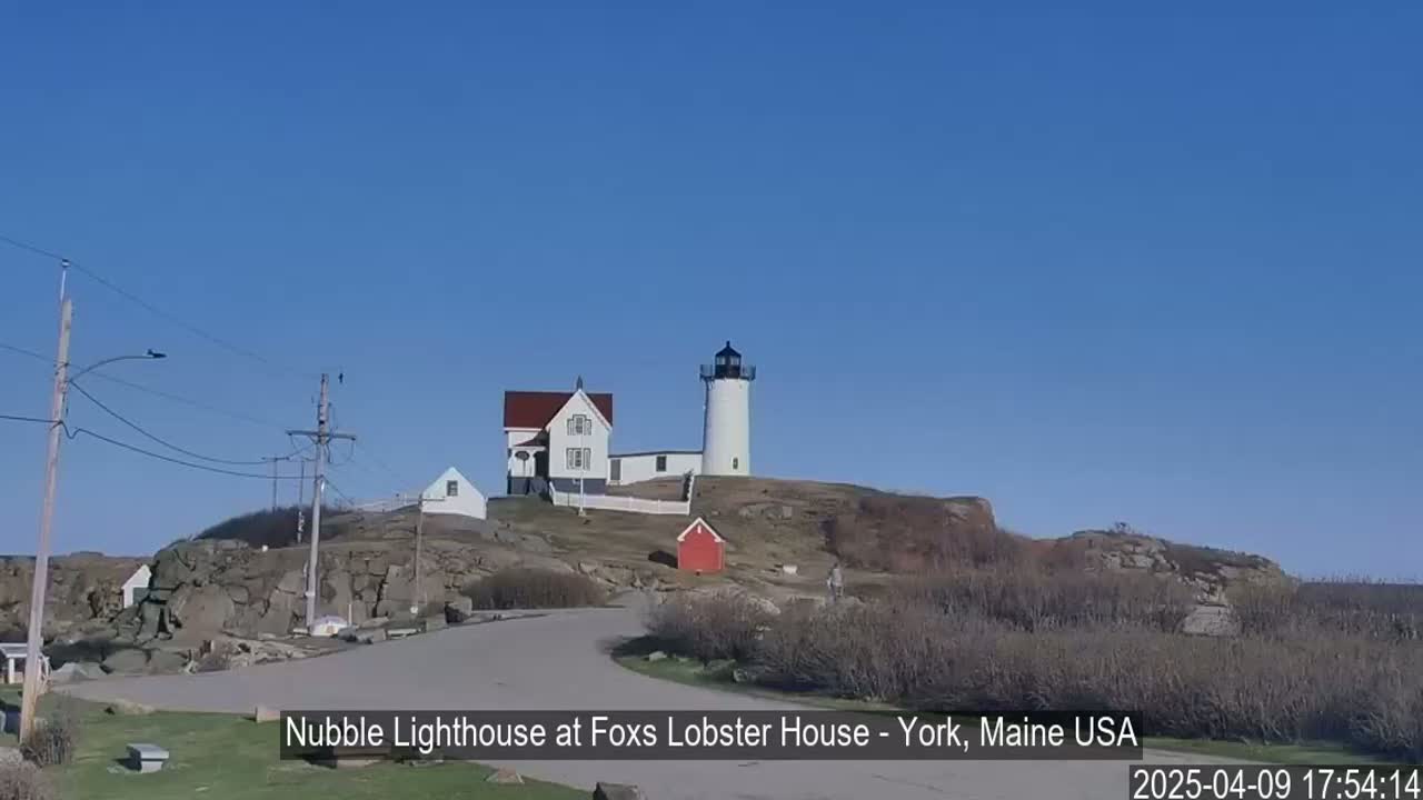 A white lighthouse and adjacent buildings sit atop a rocky hill under a clear blue sky.