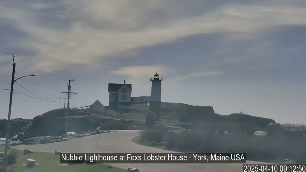 A lighthouse and adjacent building sit atop a rocky hill under a mostly cloudy sky.
