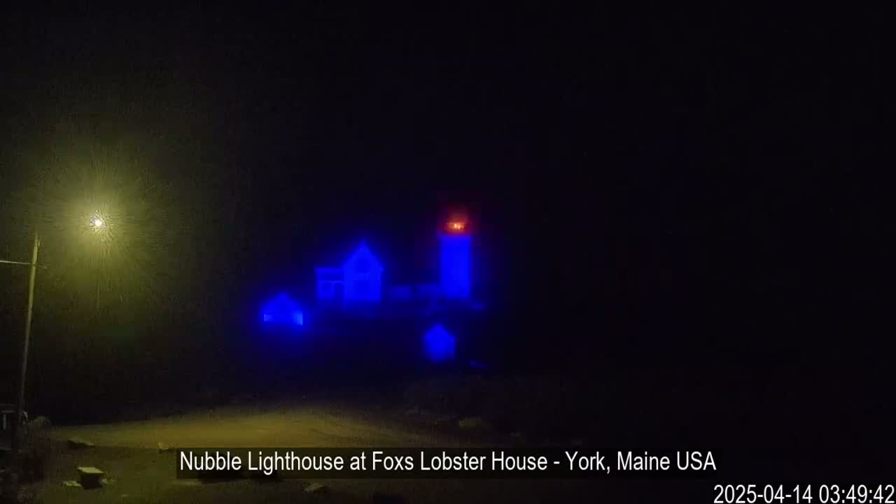 A lighthouse and nearby buildings are illuminated in blue light under foggy, nighttime conditions.