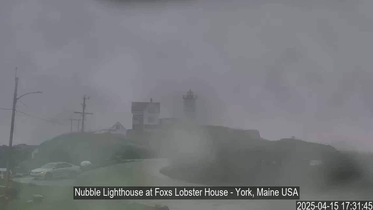 A lighthouse and other buildings are barely visible through heavy fog on a coastal road.