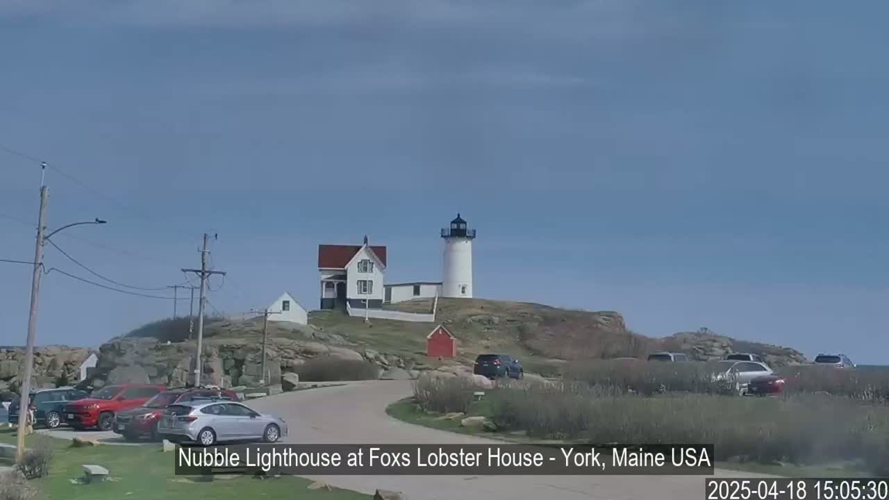 A white lighthouse stands on a rocky hill overlooking a parking lot with several cars, under a mostly clear blue sky.