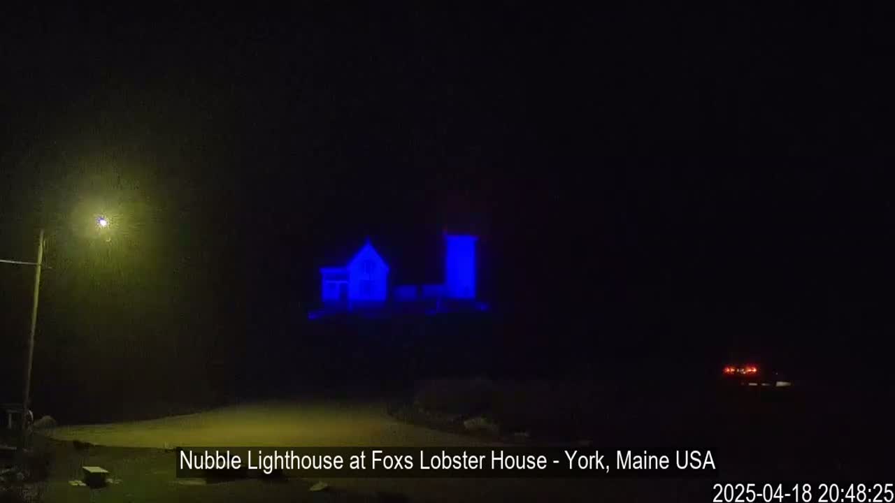 A lighthouse and keeper's house, illuminated in blue light, are visible at night in foggy conditions.