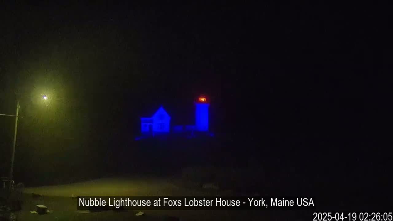 A lighthouse and a small building are illuminated in blue under a foggy, dark night.