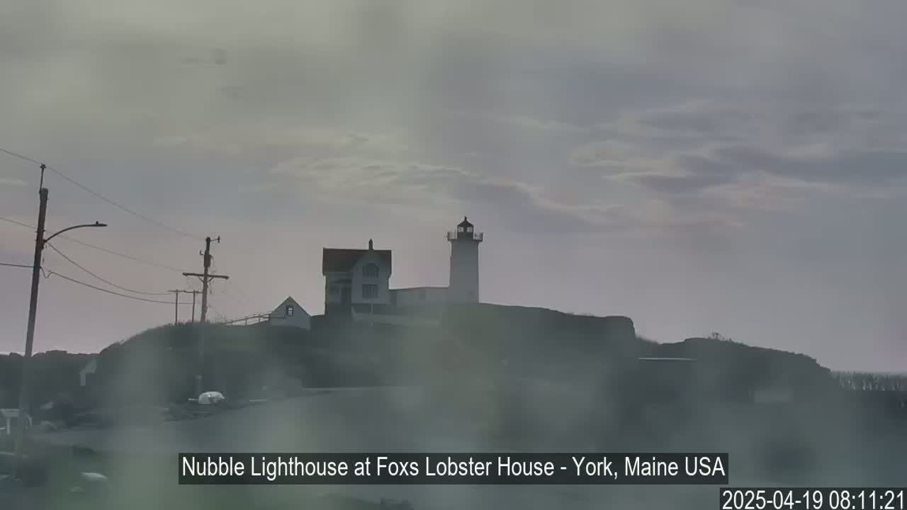 A lighthouse and a house sit on a hill shrouded in fog under a cloudy sky.