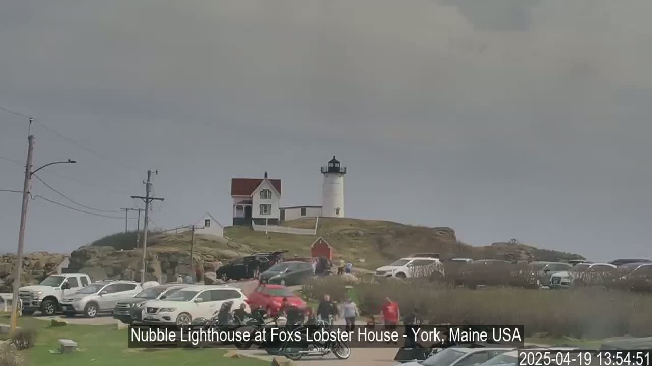 A lighthouse and several buildings sit atop a rocky hill overlooking a parking lot full of cars under an overcast sky.