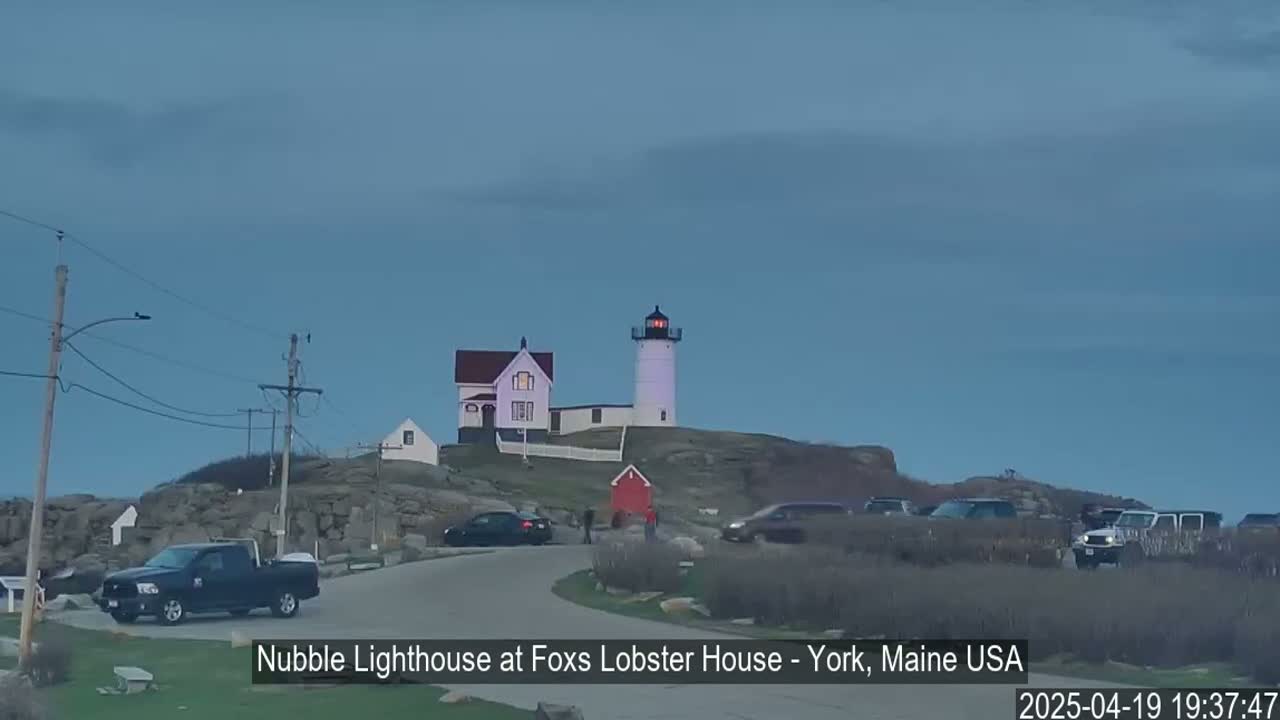 A white lighthouse and adjacent buildings sit atop a rocky hill overlooking a parking lot with several vehicles under a twilight sky.