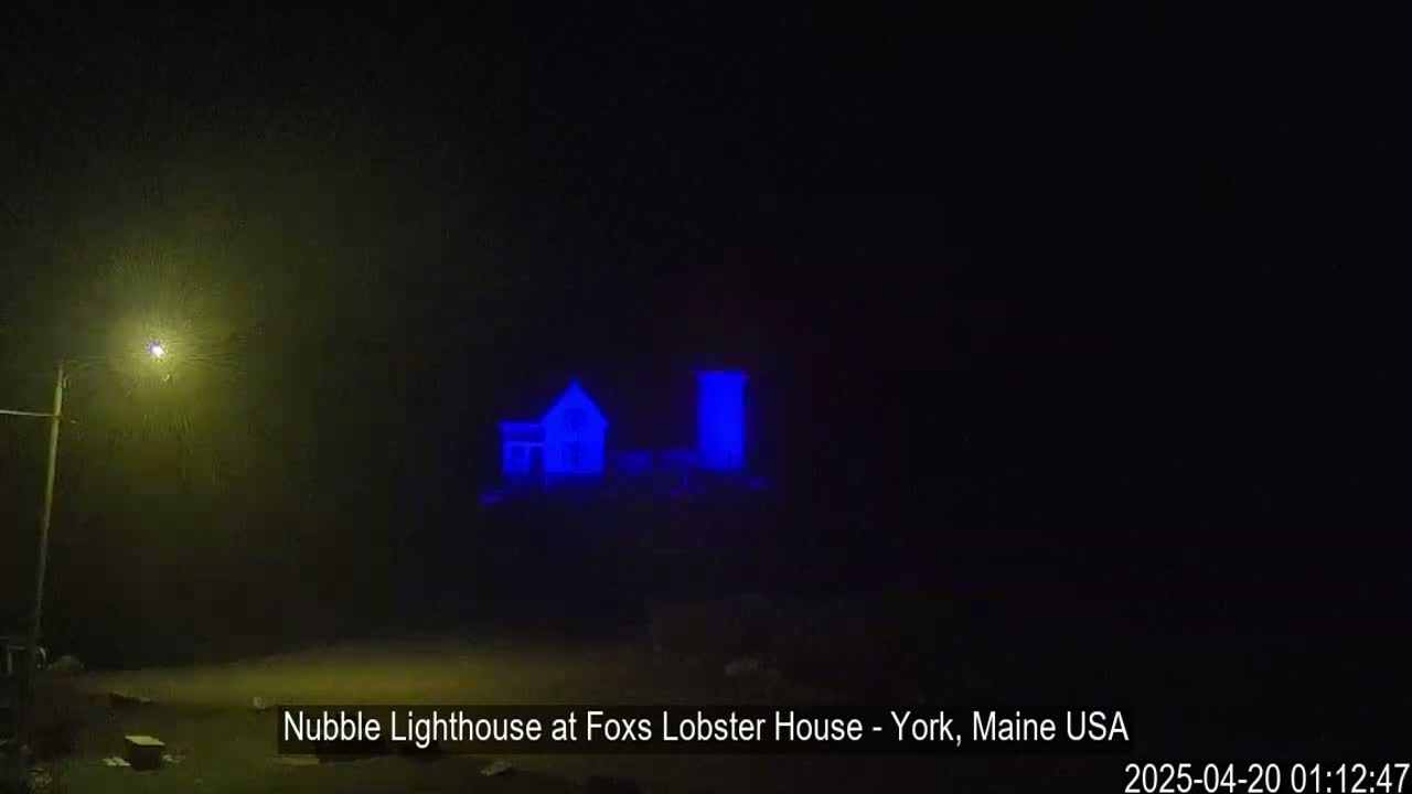 A lighthouse and a smaller building are illuminated in blue at night in foggy conditions.