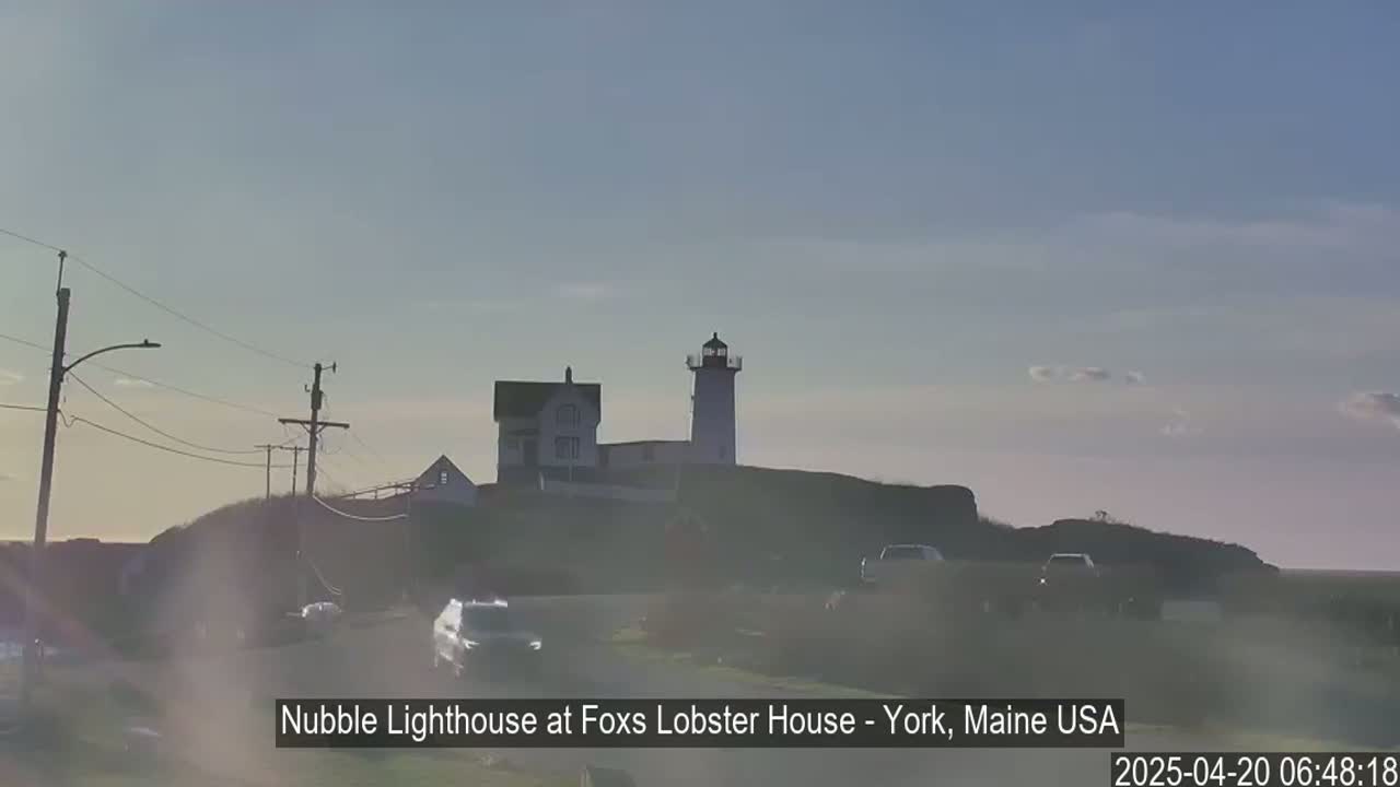 A lighthouse and adjacent building sit atop a hill overlooking a road with cars, under a mostly clear sky.