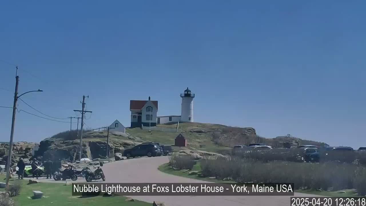 A white lighthouse and keeper's house sit atop a rocky hill overlooking a parking lot filled with cars on a sunny day.