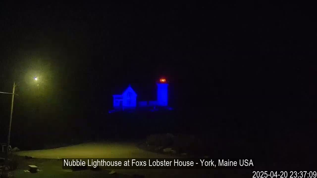 A lighthouse and keeper's house are illuminated in blue at night in foggy conditions.