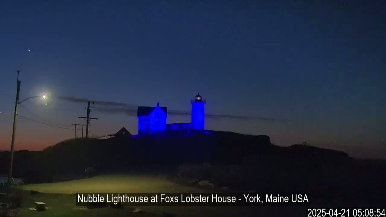 A lighthouse and a smaller building, illuminated in blue, sit atop a dark hill under a twilight sky.