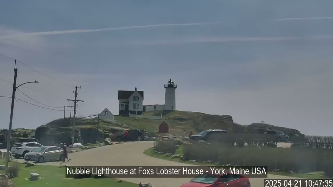 A lighthouse and a smaller white house sit atop a rocky hill on a partly sunny day, with several cars parked nearby.