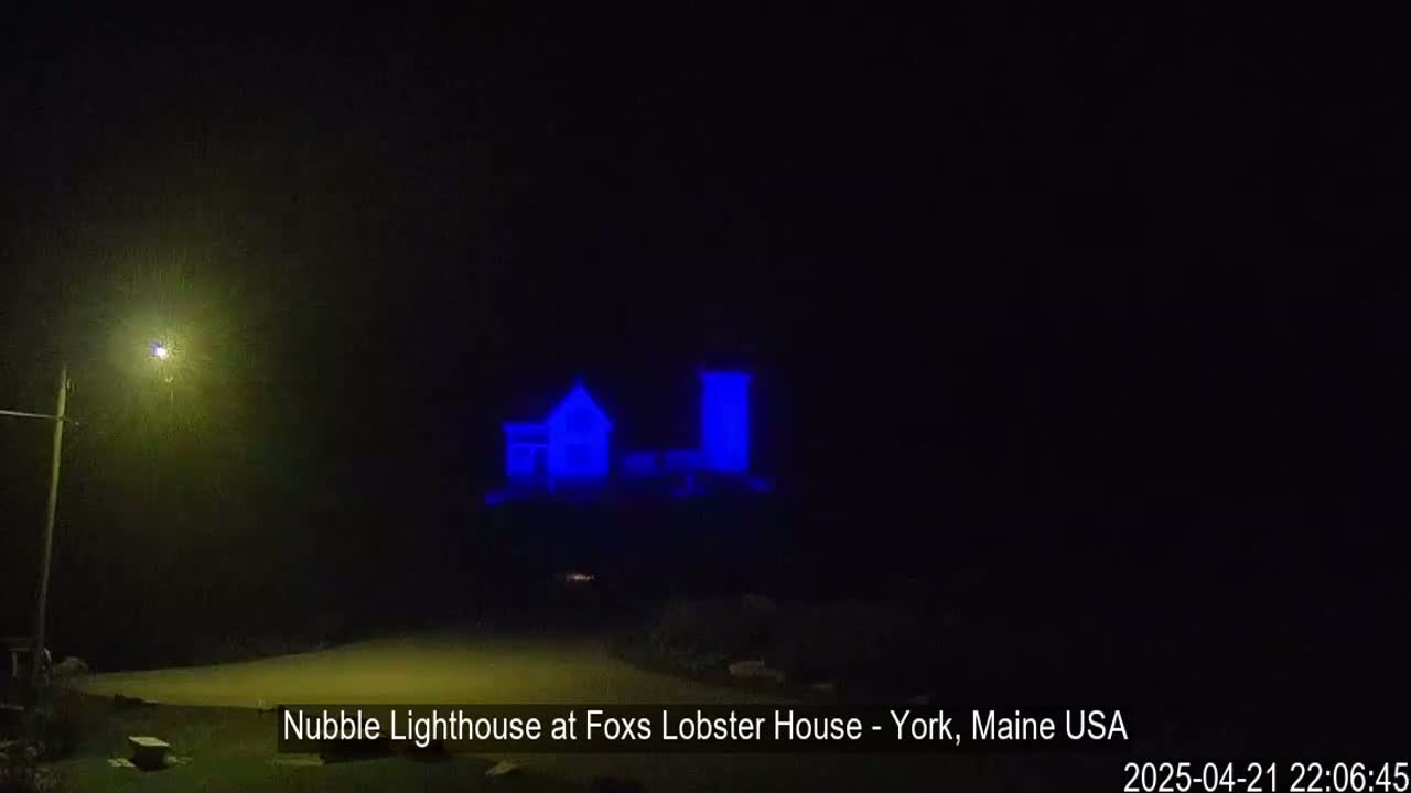 A lighthouse and adjacent building are illuminated in blue at night in hazy conditions.