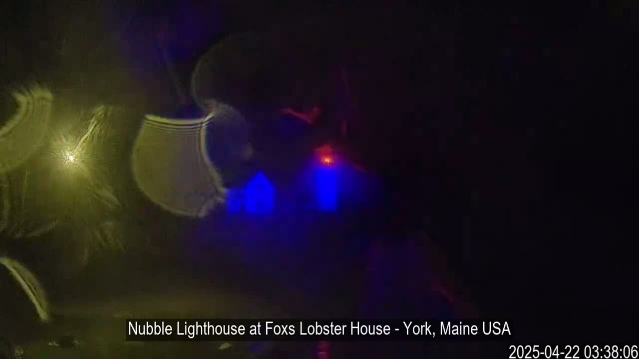 A blurry, nighttime image shows a lighthouse with blue and red lights reflecting on dark water.