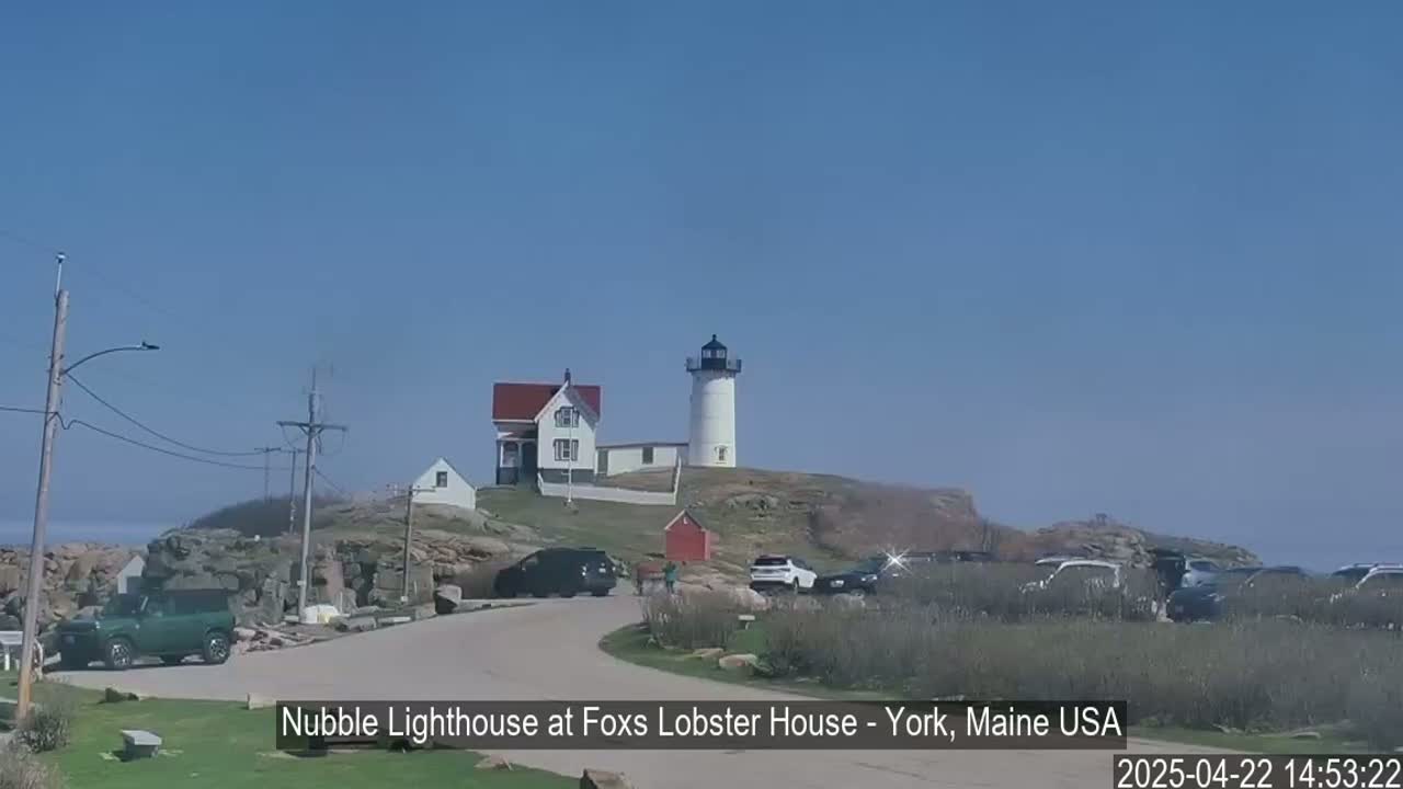 A lighthouse and several buildings sit atop a rocky outcrop overlooking a calm ocean under a clear blue sky.