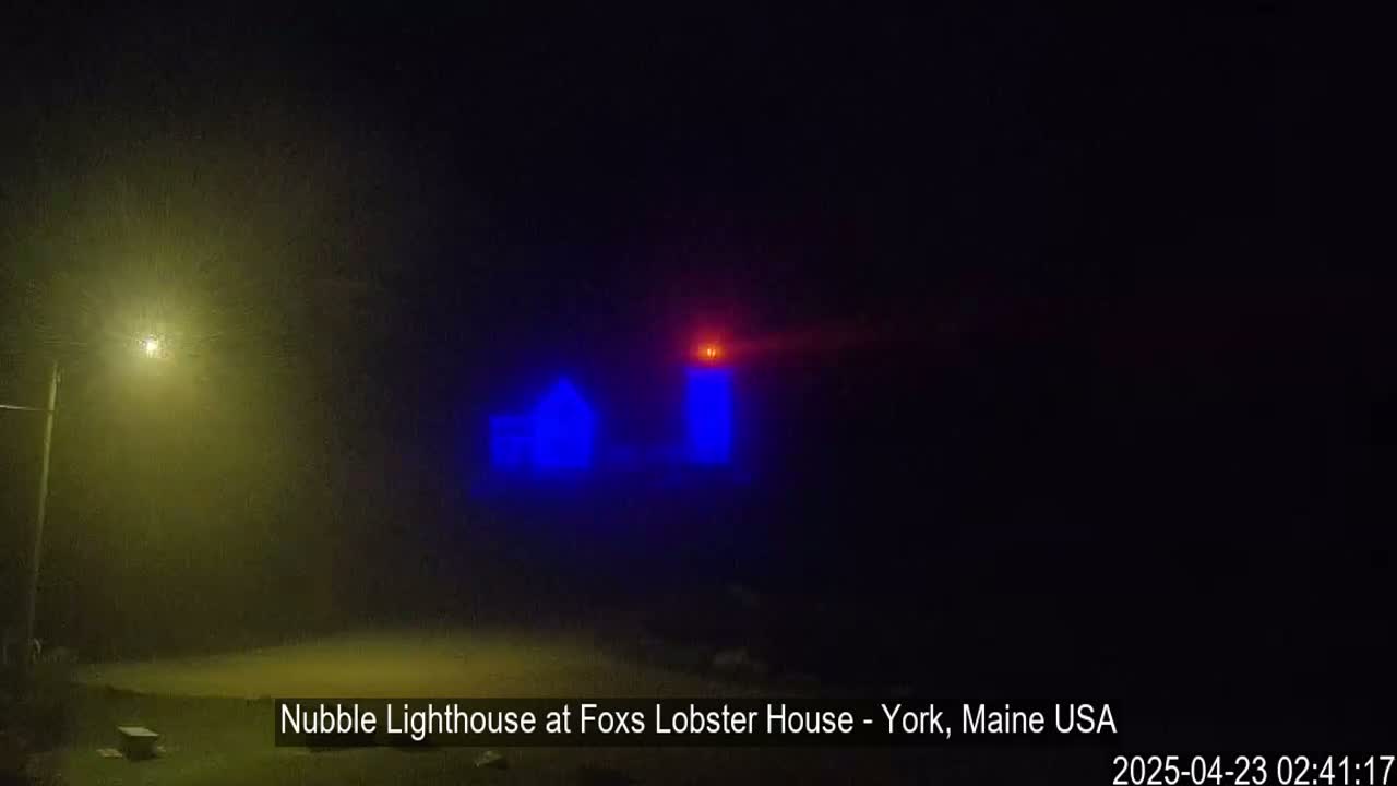 A lighthouse, illuminated in blue, is visible through a heavy fog at night.