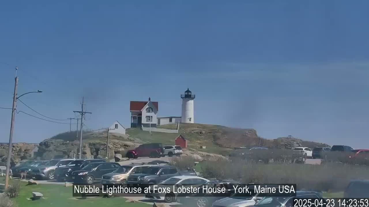 A white lighthouse sits atop a rocky hill overlooking a parking lot filled with cars under a clear blue sky.