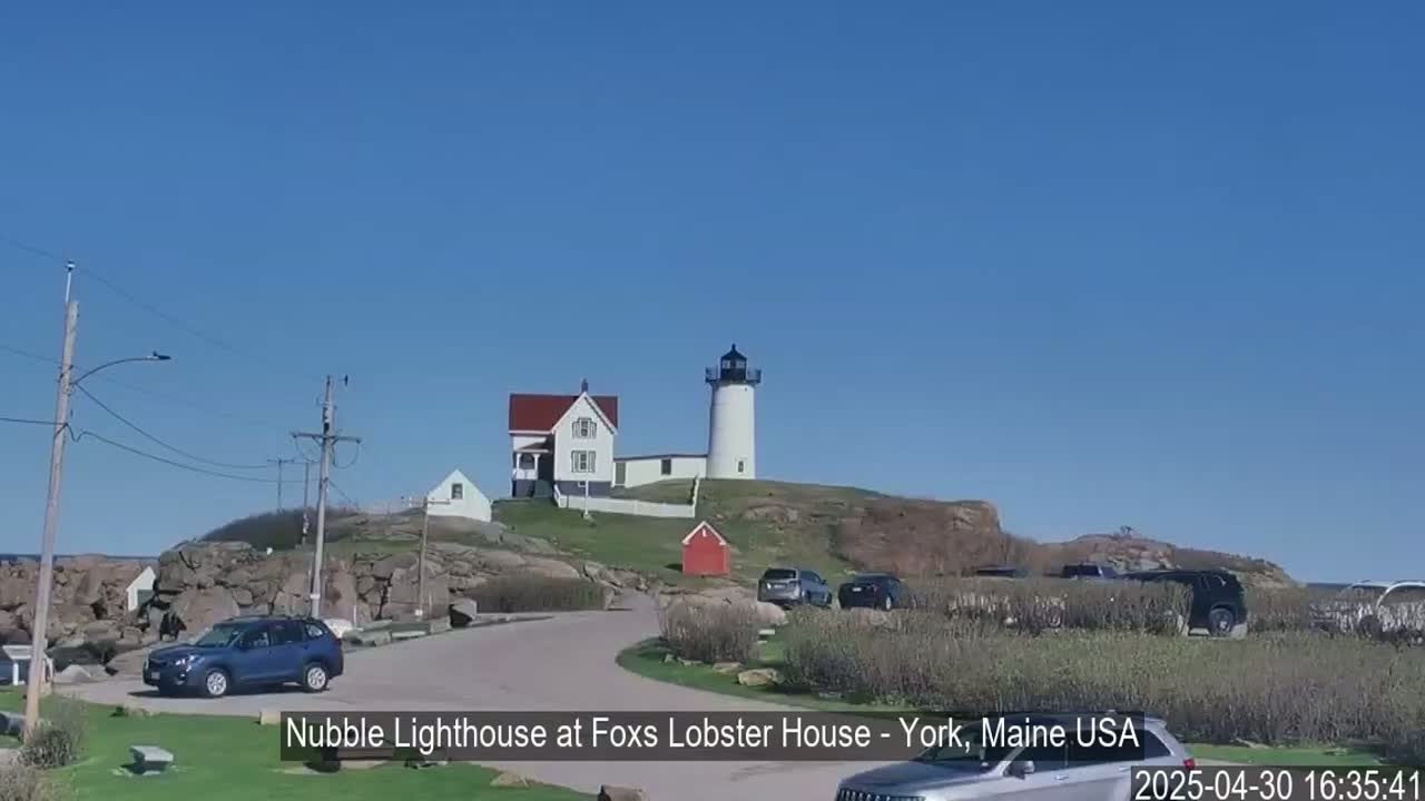 A white lighthouse sits atop a rocky hill overlooking a parking lot with several cars under a clear blue sky.
