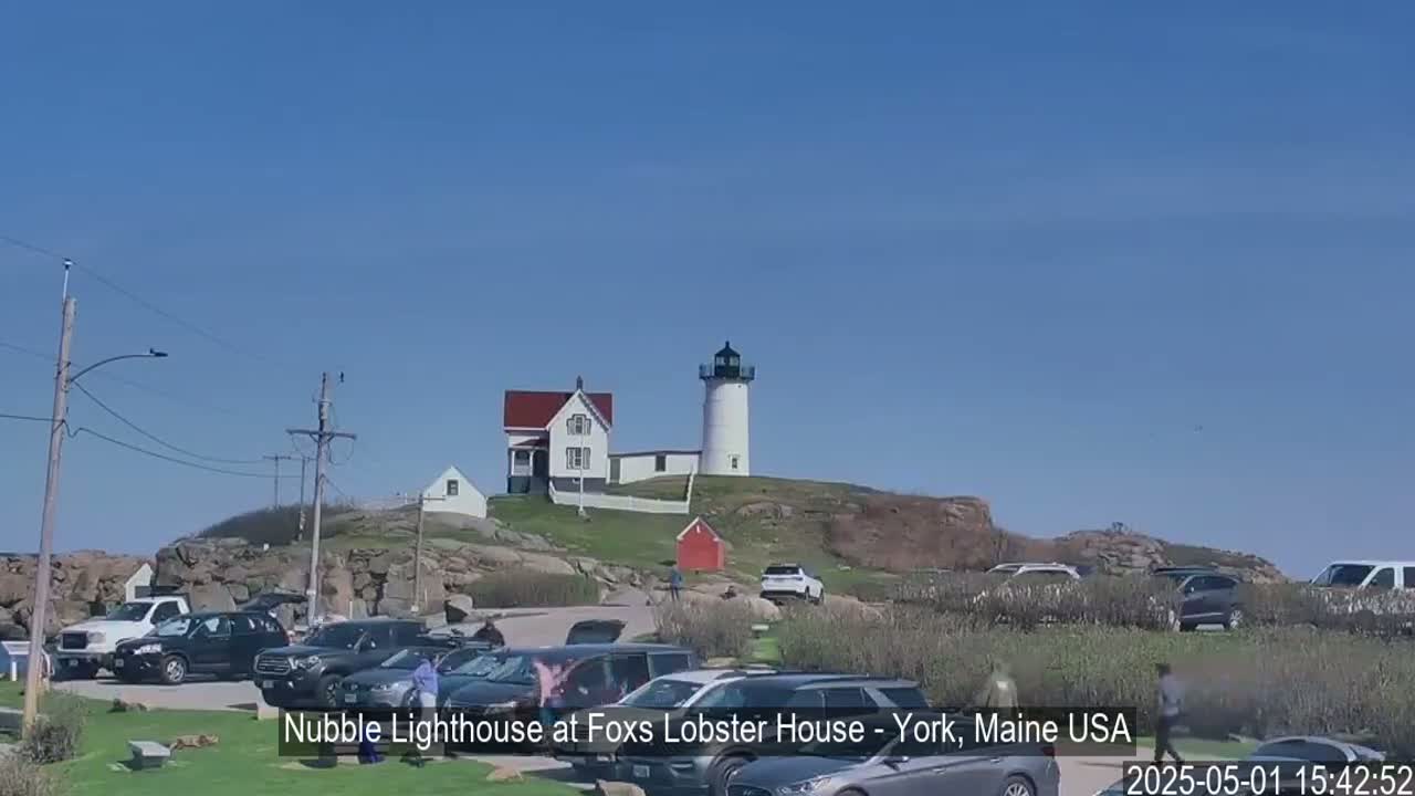 A white lighthouse sits atop a rocky hill overlooking a parking lot filled with cars, under a clear blue sky.