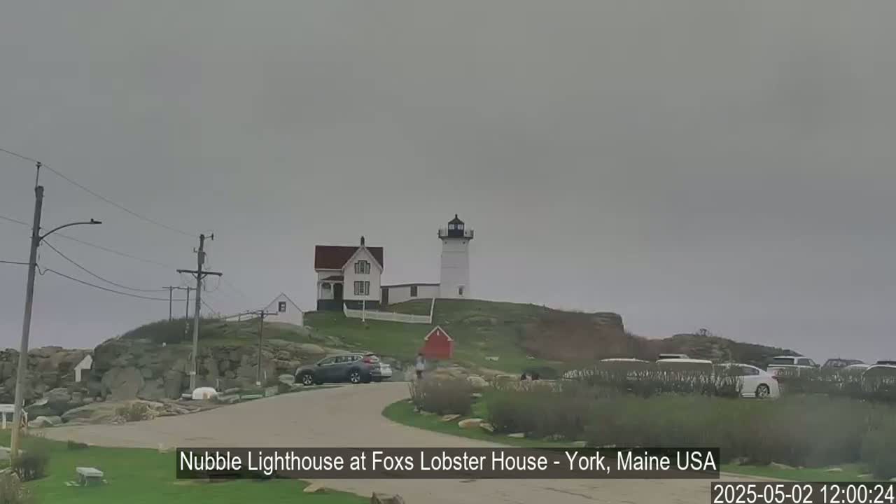 A white lighthouse and a smaller white building with a red roof sit atop a rocky hill under an overcast sky, with several cars parked in a lot nearby.