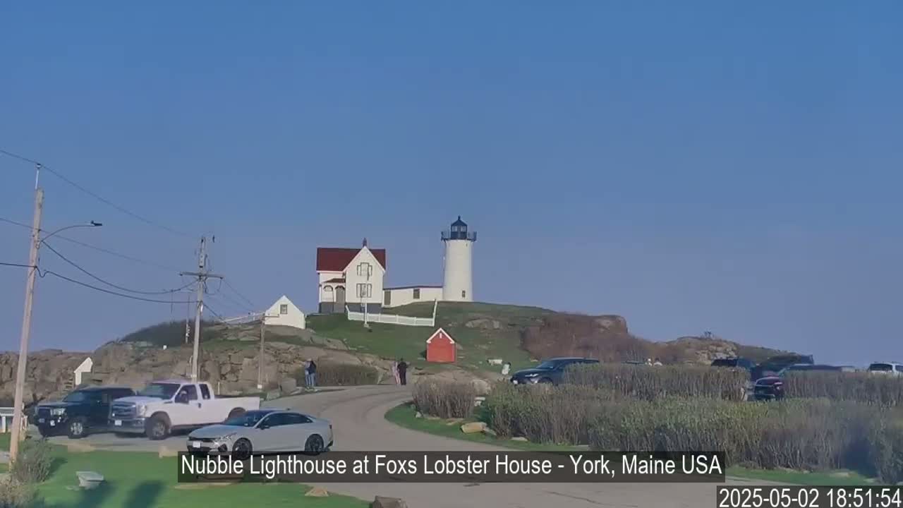 A white lighthouse sits atop a rocky hill overlooking a parking lot filled with cars on a clear, sunny day.
