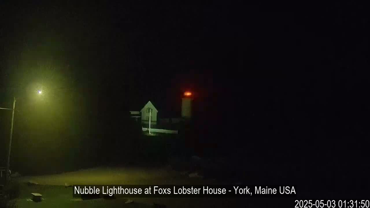 A lighthouse with a red light shines brightly in the foggy darkness, illuminated by a nearby streetlight.