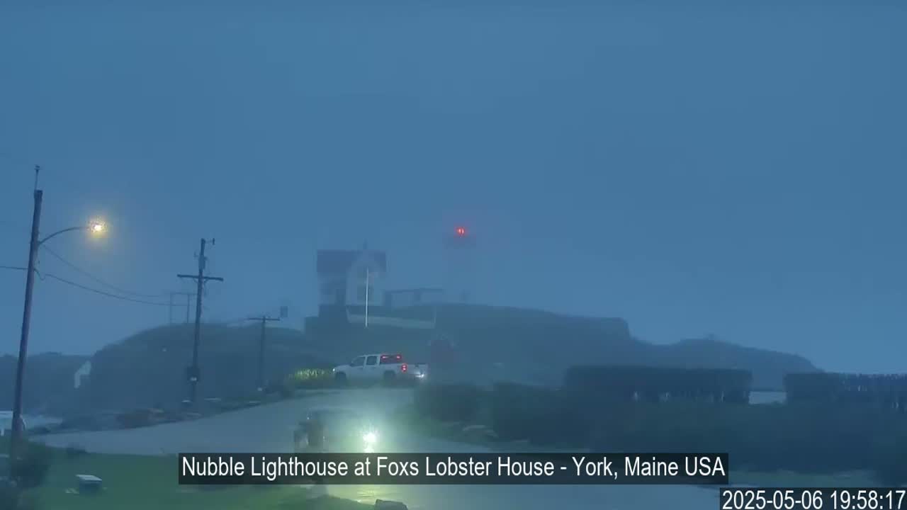 A lighthouse is visible through a hazy, dark blue sky, with a vehicle parked nearby on a dimly lit road.