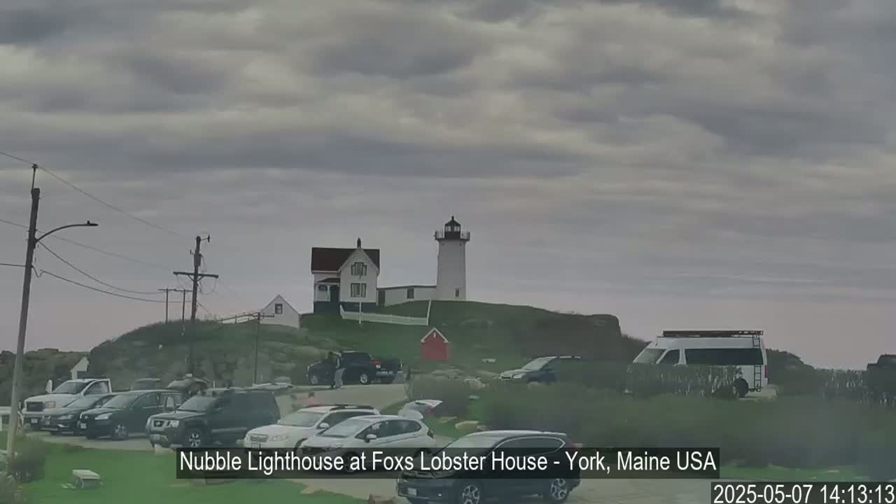A lighthouse and accompanying building sit atop a grassy hill overlooking a parking lot filled with cars under an overcast sky.