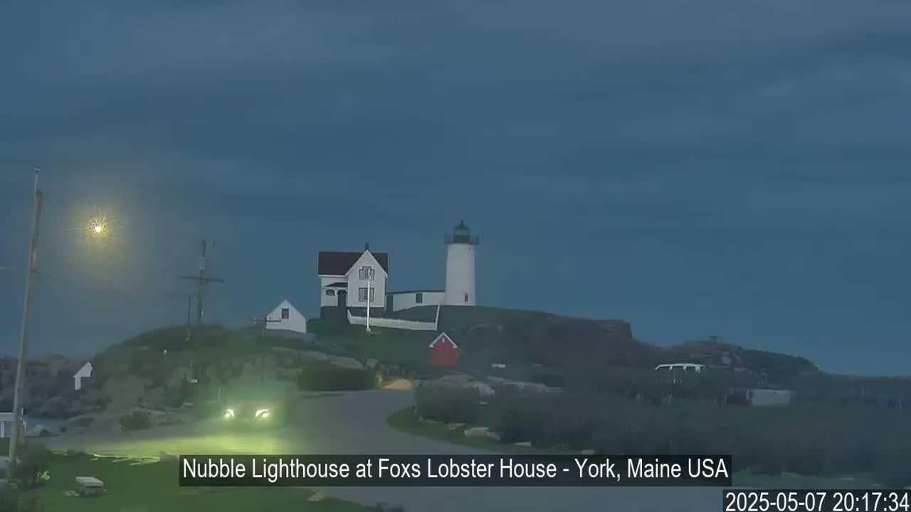 A white lighthouse and accompanying buildings sit on a rocky hill under a dusky, overcast sky.