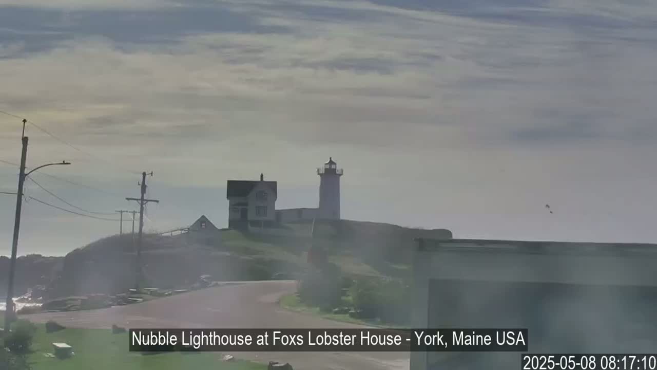 A lighthouse and a smaller building sit atop a hazy, rocky outcropping overlooking a calm ocean under a mostly cloudy sky.