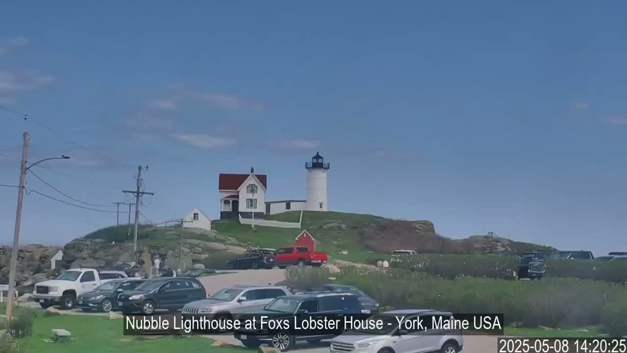 A white lighthouse with a red-roofed building sits atop a rocky hill overlooking a parking lot filled with cars on a sunny day.
