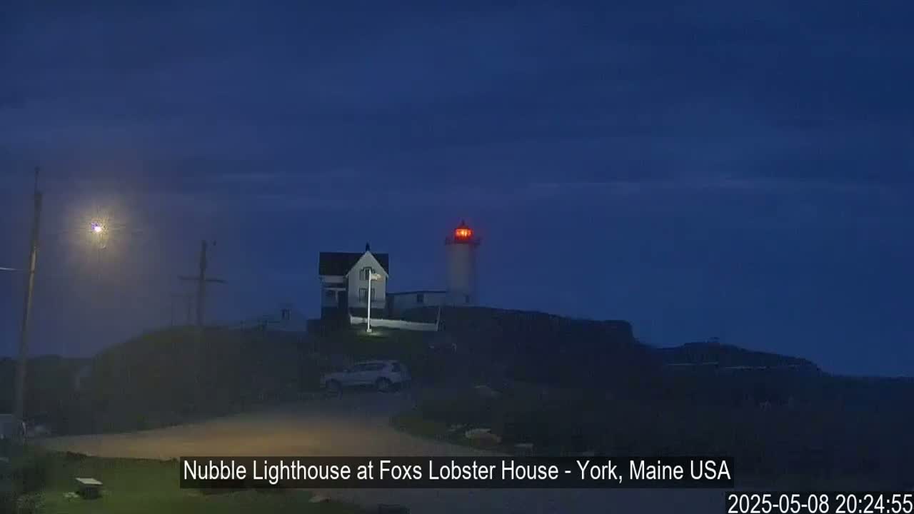 A lighthouse with a red light atop a dark hill is visible at night under a hazy, dark blue sky.