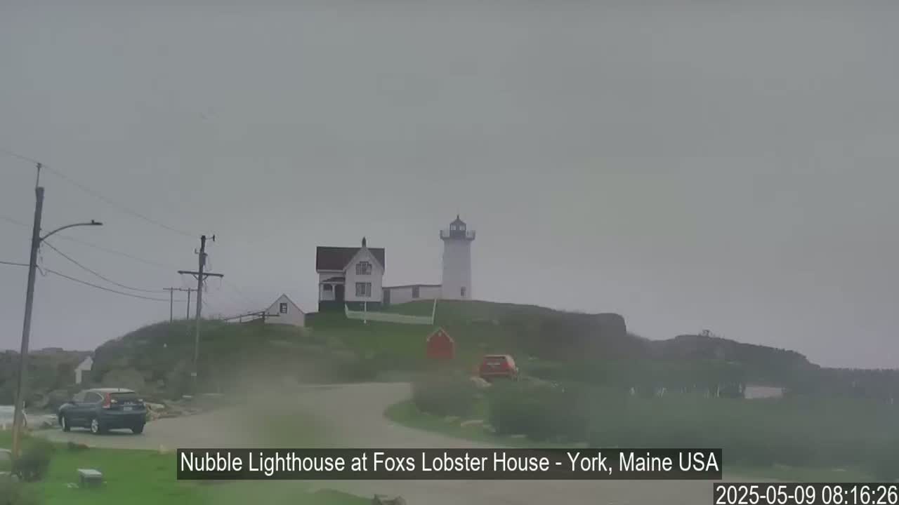 A lighthouse and other buildings sit atop a hill on an overcast day, with a car parked nearby.