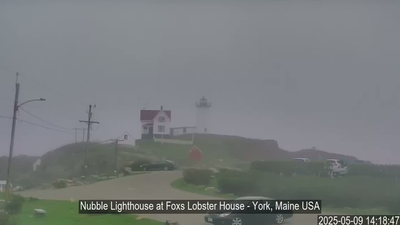 A lighthouse and a smaller building are barely visible through a thick fog on a hillside, with cars parked nearby.
