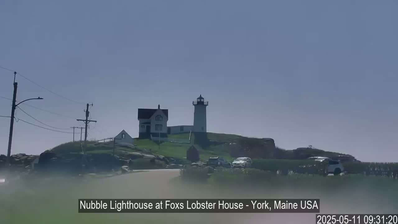 A lighthouse and a small house sit atop a rocky hill on a hazy, sunny day with several cars parked nearby.