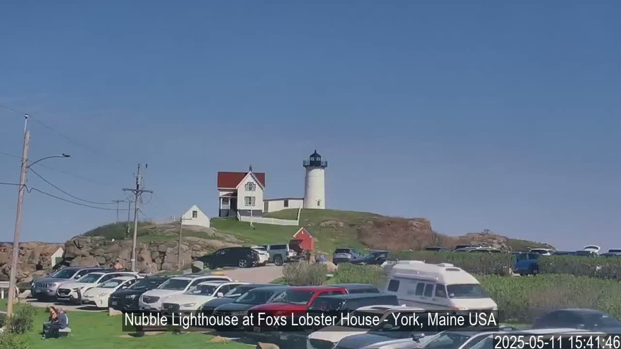 A lighthouse sits atop a rocky hill overlooking a large parking lot filled with cars and an RV, under a clear blue sky.