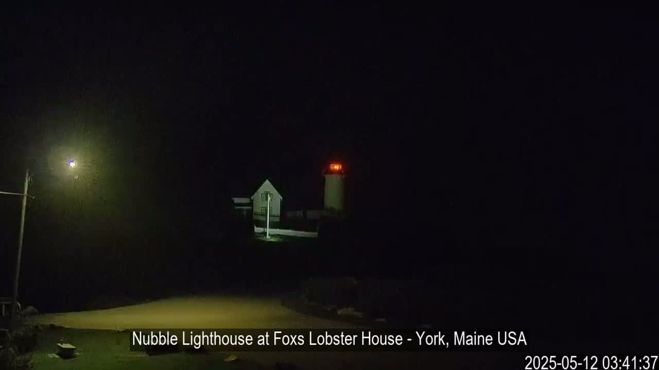 A lighthouse with a red light illuminates the night, next to a smaller building.