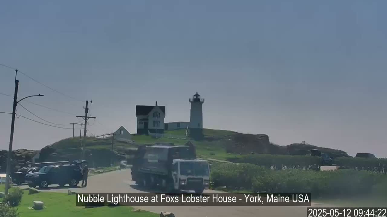 A lighthouse and a smaller building sit atop a grassy hill, with a garbage truck and several cars parked nearby on a clear day.
