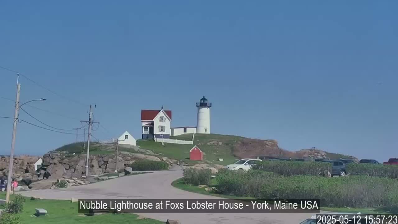 A white lighthouse and accompanying buildings sit atop a rocky hill overlooking a paved road and a parking lot on a clear, sunny day.