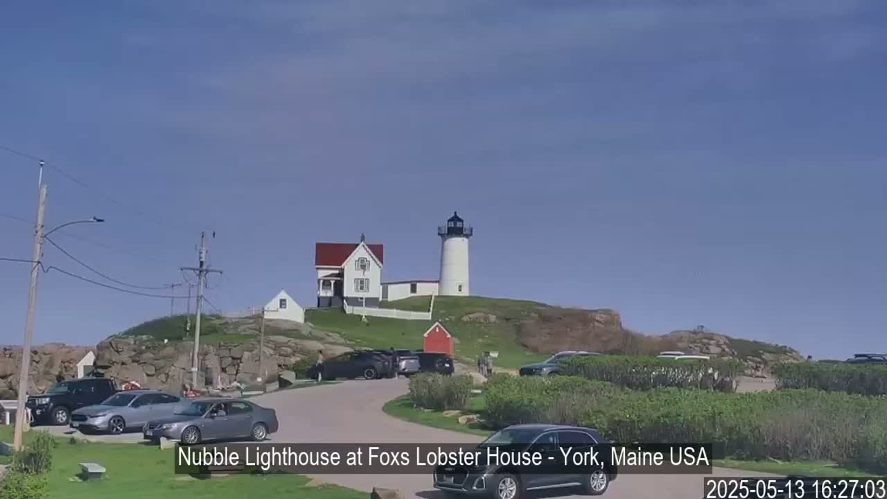 A lighthouse and accompanying buildings sit atop a rocky hill under a clear blue sky, with several cars parked in a nearby lot.