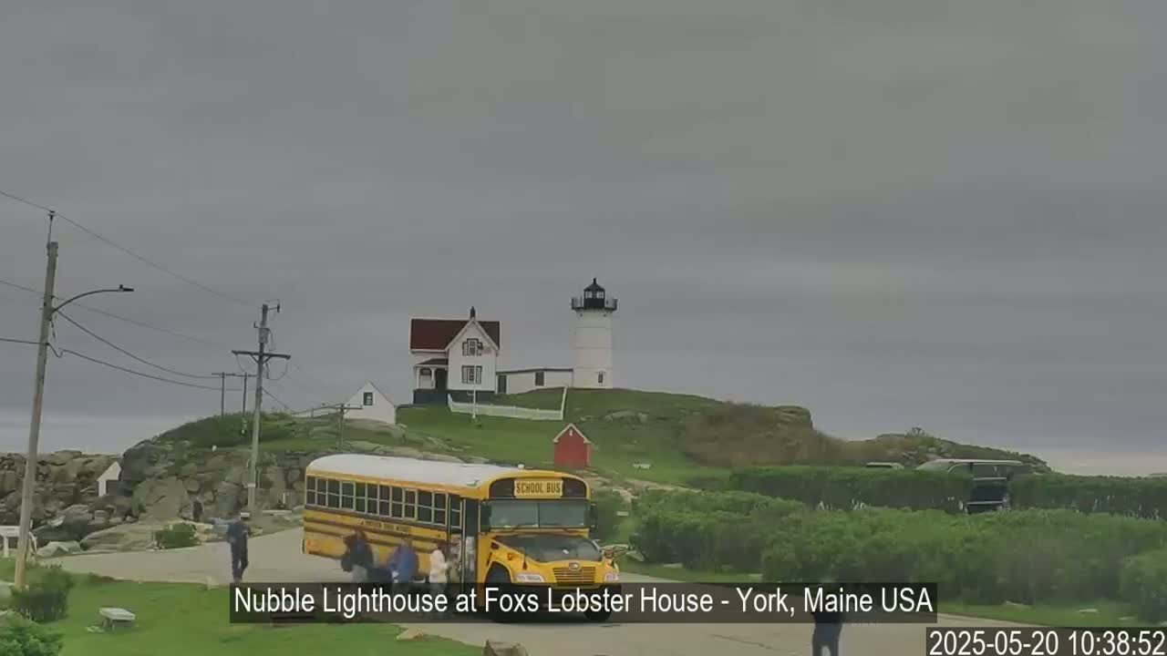 A lighthouse and keeper's house sit atop a rocky hill under an overcast sky, with a school bus and several people near the base.