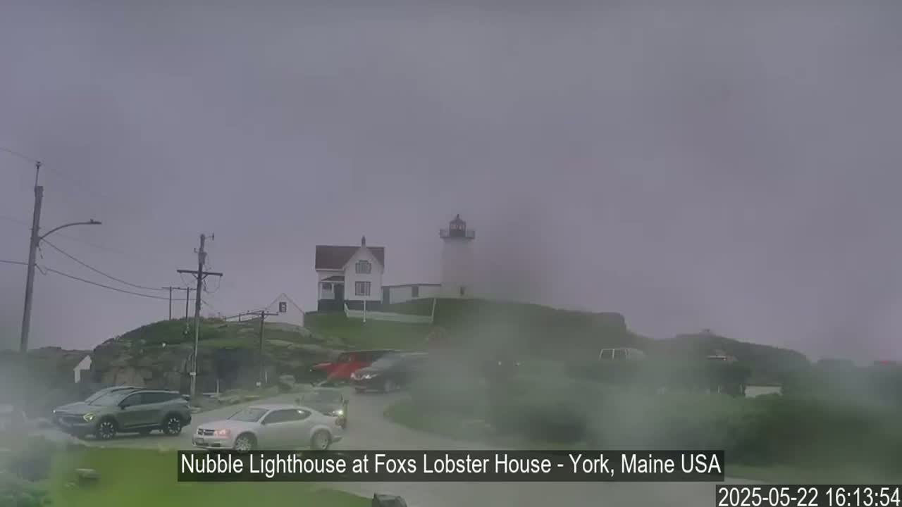 A lighthouse and several cars are visible through a heavy fog on a cloudy day.