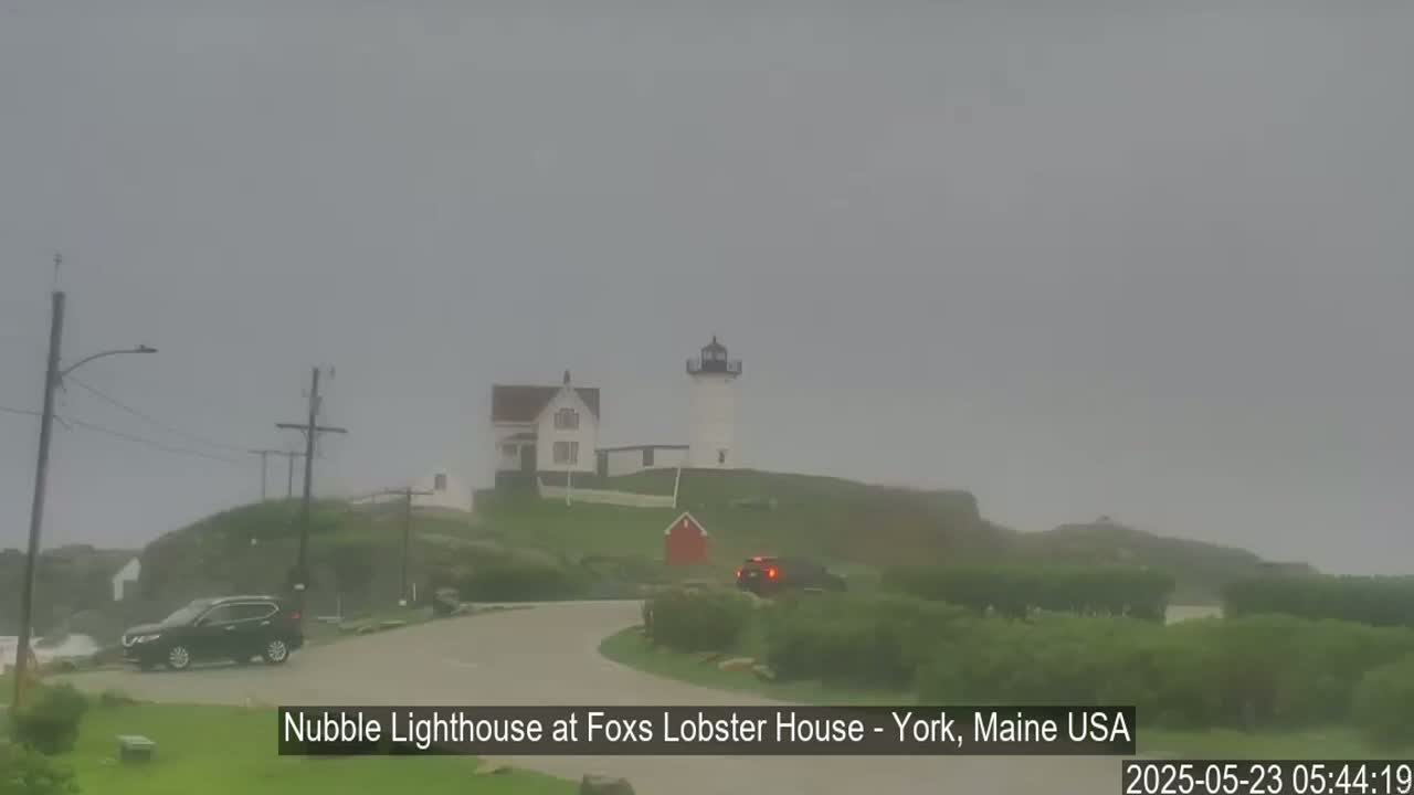 A lighthouse and accompanying buildings sit on a grassy hill overlooking a road on a foggy day.