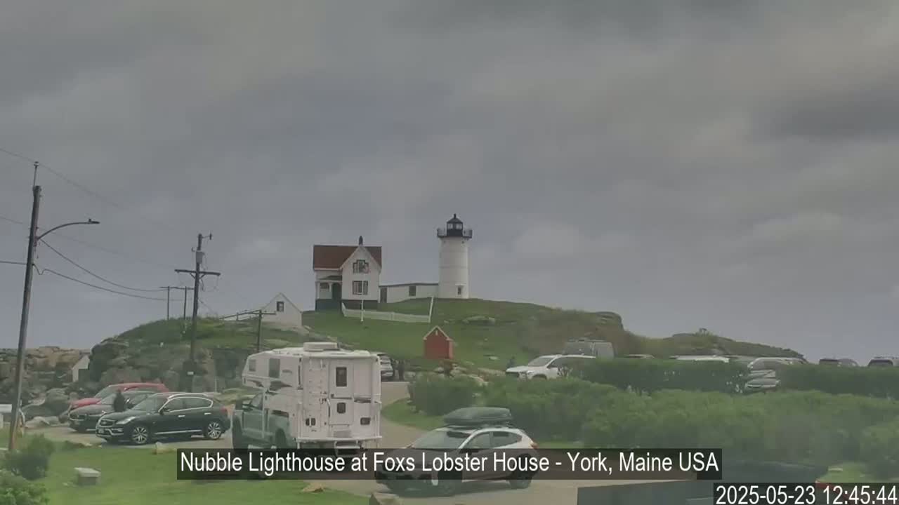 A lighthouse and several cars are parked on a grassy hill under an overcast sky.