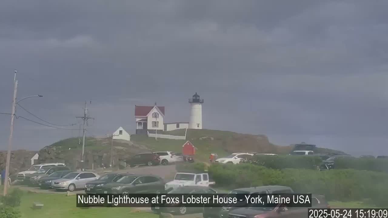 A white lighthouse sits atop a hill with several cars parked in a lot below, under an overcast sky.