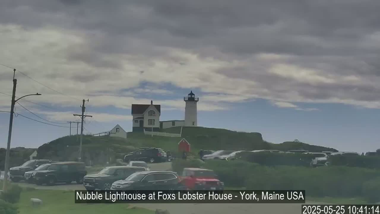 A lighthouse and a smaller building sit atop a grassy hill overlooking a parking lot full of cars under an overcast sky.